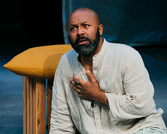 Pericles in shabby white shirt sits on the floor next to a cushioned stool as he holds his hand to his breast as a tear runs down his left cheek. 